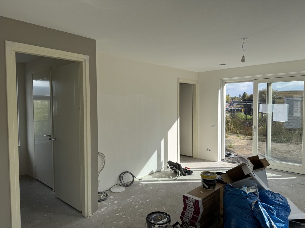 Woonkamer van een net opgeleverd huis met kale wanden, dozen laminaat en emmers op de betonvloer