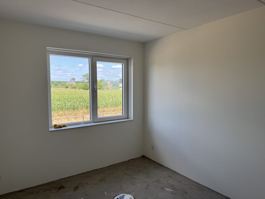 Lege slaapkamer met lichte, strakke wanden en een raam dat uitkijkt op een maisveld