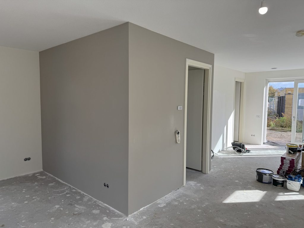 Grijs gesausde muur rond een meterkast in een woonkamer, met verfemmers bij de tuindeur